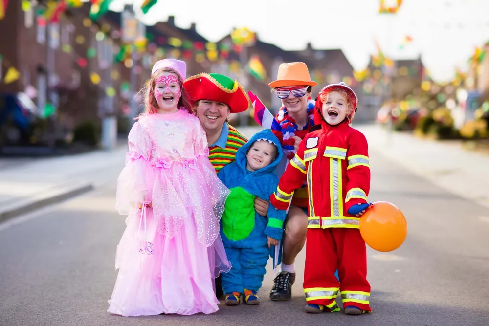 Group of five children in colorful costumes and face paint, smiling on a decorated street during a festive event.