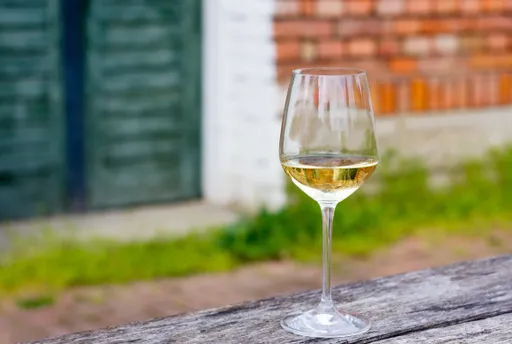 Ein Glas Weißwein auf einem rustikalen Holztisch im Freien, im Hintergrund eine verschwommene Backsteinmauer und Grünpflanzen.