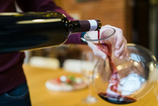 Rotwein wird aus einer Flasche in eine Glaskaraffe auf einem Holztisch mit unscharfem Hintergrund gegossen.