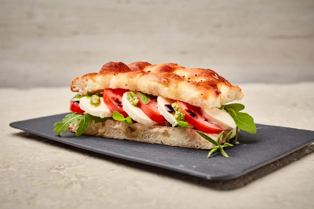 Caprese-Sandwich mit frischem Mozzarella, Tomaten, Rucola und Pesto auf Focaccia-Brot, serviert auf einem schwarzen Schieferbrett.