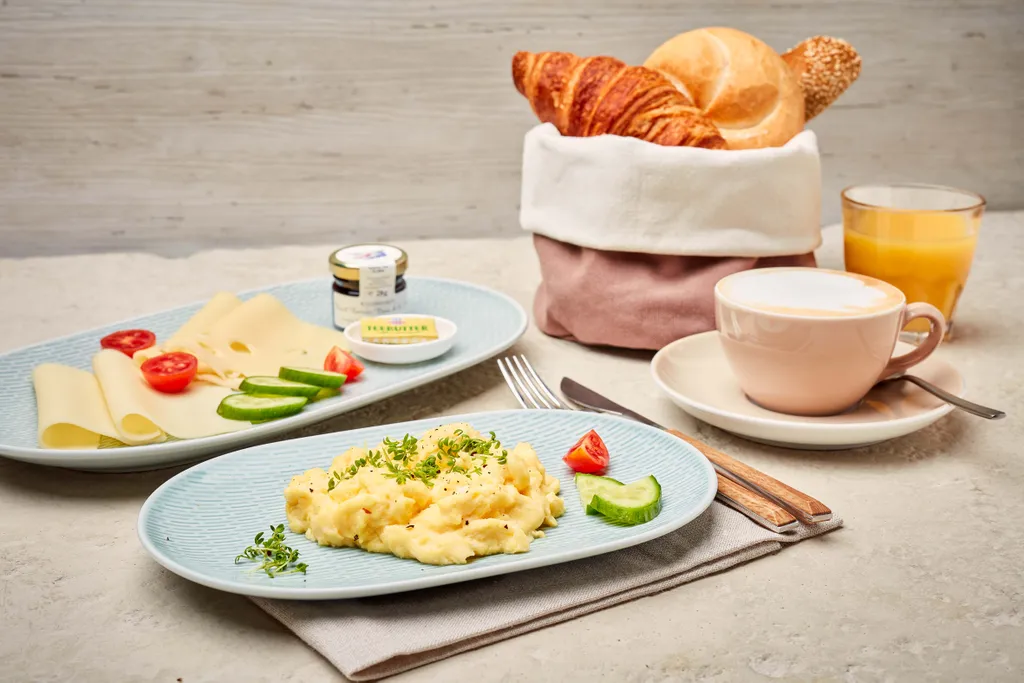 Ein Frühstücksbuffet mit Rührei, Käse, Tomaten, Gurken, Croissants, einem Brötchen, Orangensaft und einer Tasse Cappuccino auf einem hellen Tisch.
