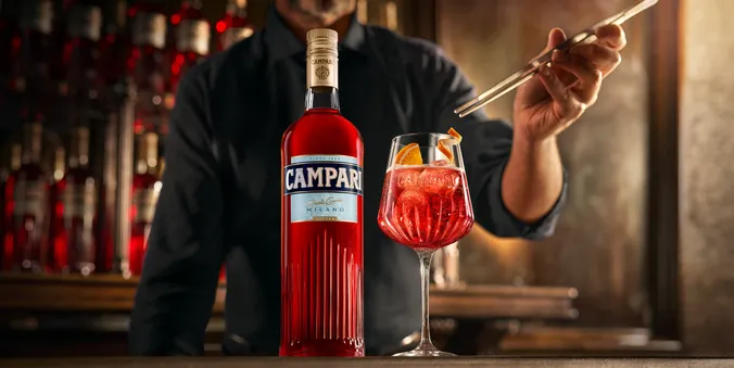 A bottle of Campari beside a glass with a red cocktail and orange garnish, against a blurred bar background.