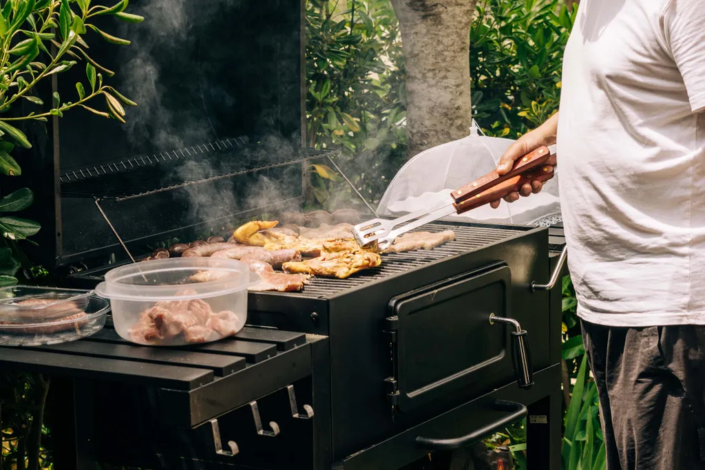 Eine Person, die auf einem rauchenden Holzkohlegrill Würstchen und Hähnchen grillt, daneben stehen Schüsseln mit Fleisch, umgeben von üppigem Grün.