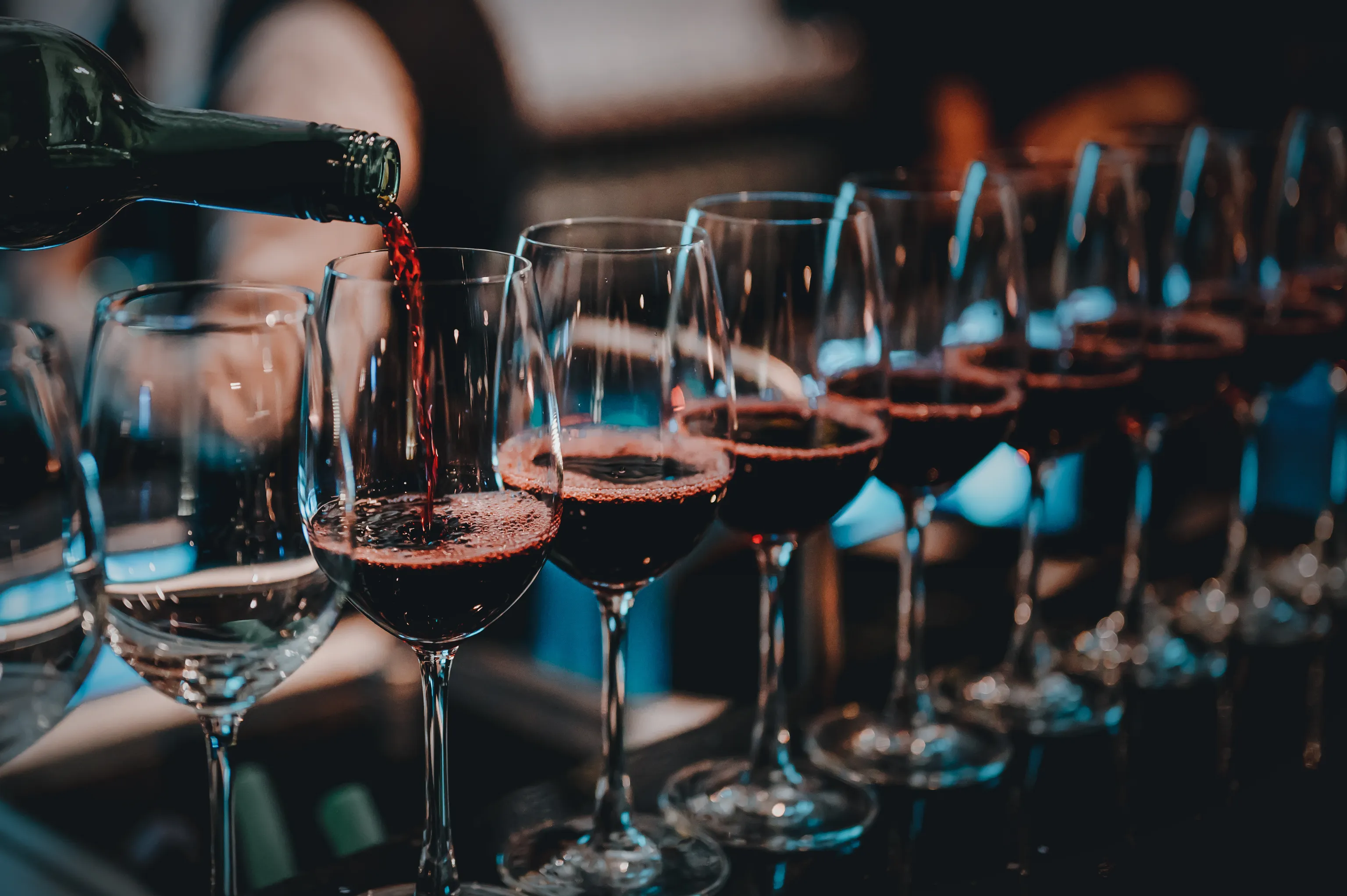 Rotwein wird in eines von mehreren nebeneinander aufgereihten Weingläsern auf einer schummrig beleuchteten Bartheke eingeschenkt.