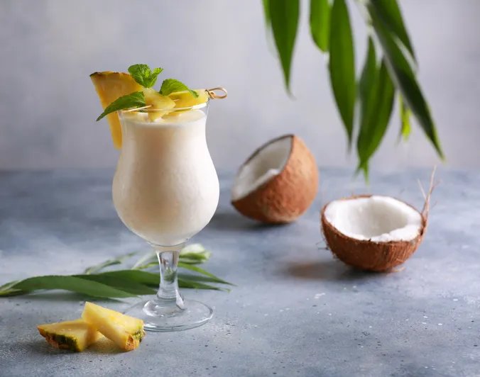 Eine eisgekühlte Piña Colada, garniert mit Ananas und Minze in einem Glas, umgeben von Kokosnussschalen, Ananasscheiben und grünen Blättern auf einem grauen Untergrund.