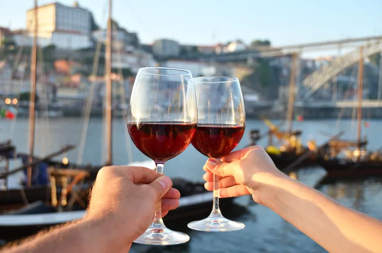 Zwei Hände, die Weingläser mit Rotwein halten und am Flussufer anstoßen, im unscharfen Hintergrund sind eine Brücke und Boote zu sehen.