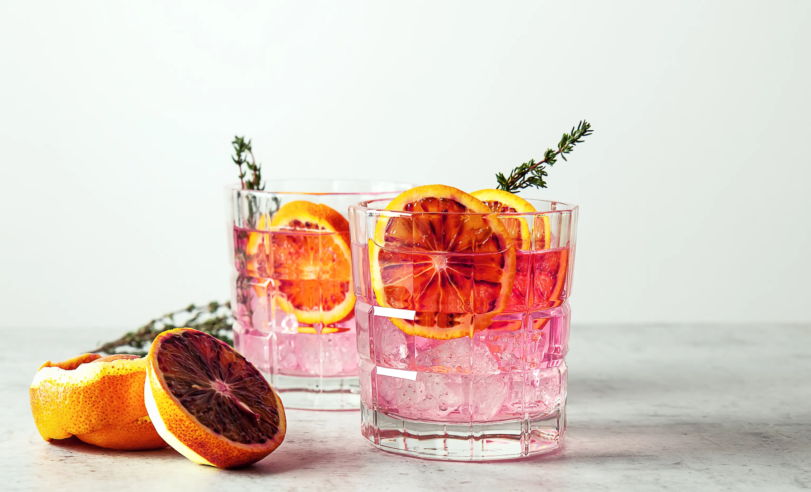 Zwei Gläser mit rosa Cocktails, garniert mit Blutorangen-Scheiben und Thymian, stehen auf einer Marmorplatte, daneben liegen frische Orangenscheiben.