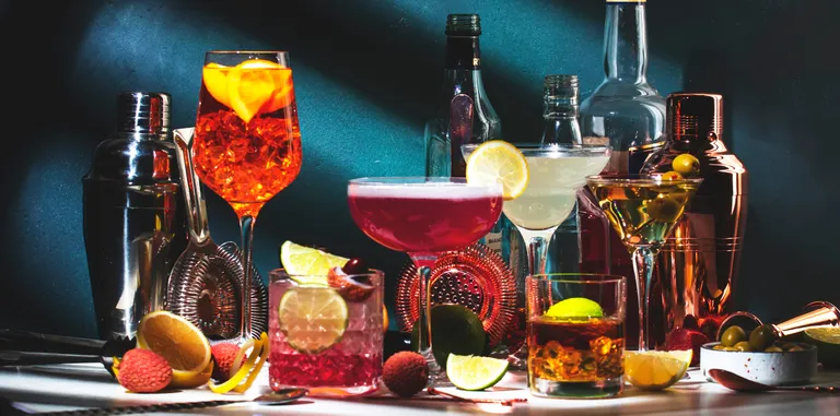 A variety of colorful cocktails in glasses with garnishes, surrounded by cocktail shakers, bottles, and sliced fruits on a dark background.