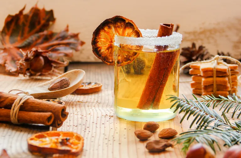Ein Glas gewürzter Punsch mit Zimtstangen und einer getrockneten Orangenscheibe auf einem Holztisch, umgeben von Nüssen, Gewürzen und Herbstlaub.