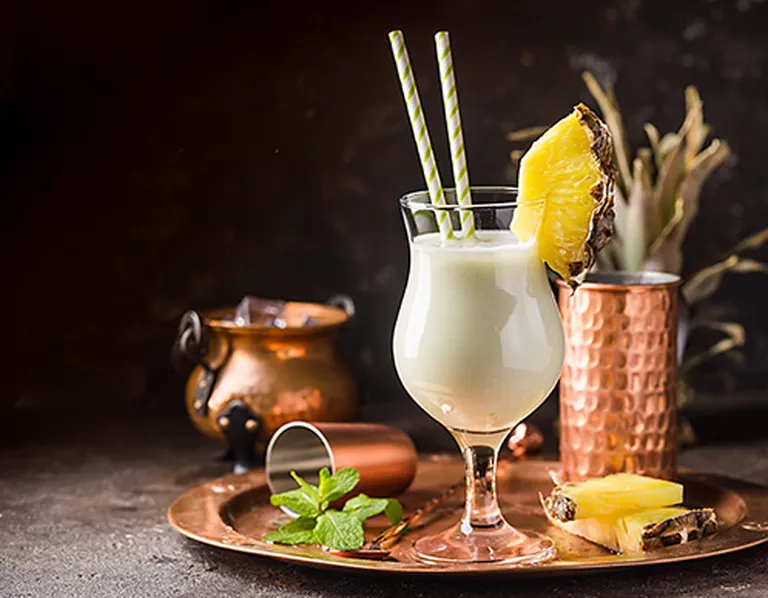 Piña Colada in einem Cocktailglas, darin zwei Strohhalme und ein Stück Ananas. Auf Messingtablet, daneben weitere Ananasstücke, sowie Minze und ein Messbecher.
