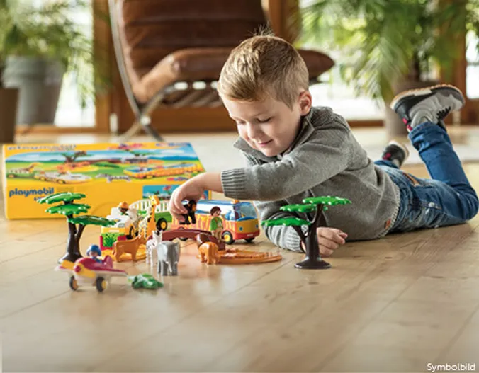 Ein kleines Kind liegt spielend auf dem Boden und vor ihm kleine Spielfigute, wie Tiere aus Afrika (Elefant, Giraffe uvm.) und Safari-Fahrzeugen (Reisebus und Offroad-Wagen)..