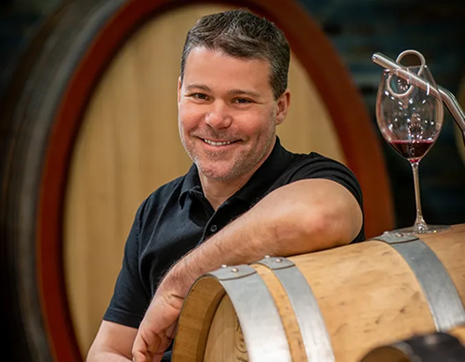 EIn Mann lehnt sich an ein Fass auf dem ein Weinglas, gefüllt mit Rotwein steht und schaut in die Kamera