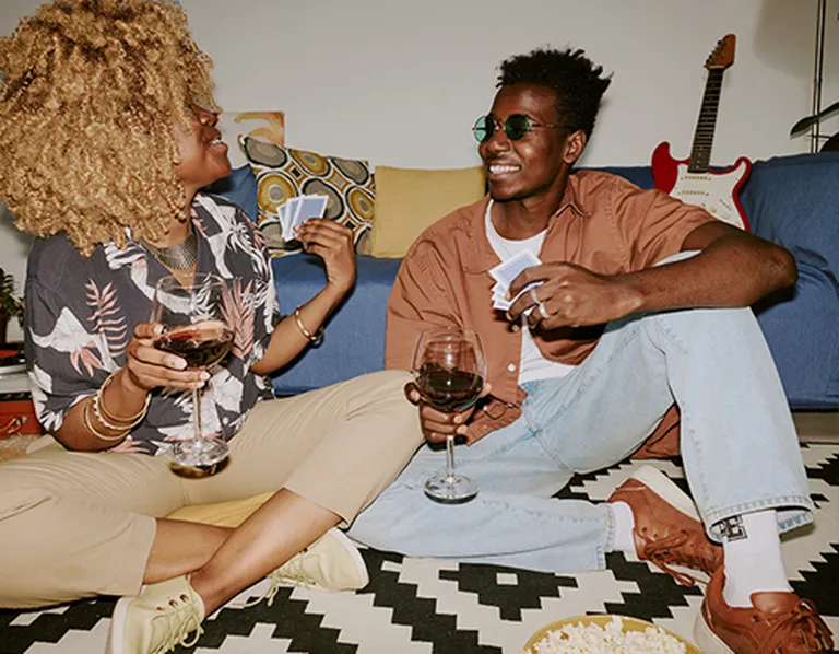 Two friends sit on a patterned rug, holding glasses of wine and playing cards. A guitar and cushions are in the background.