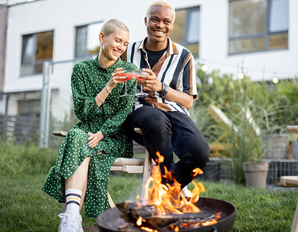 A Woman in a green dotted dress and a man in a oversized shirt and black pants sitting by a campfire, laughing, holding a glass of wine