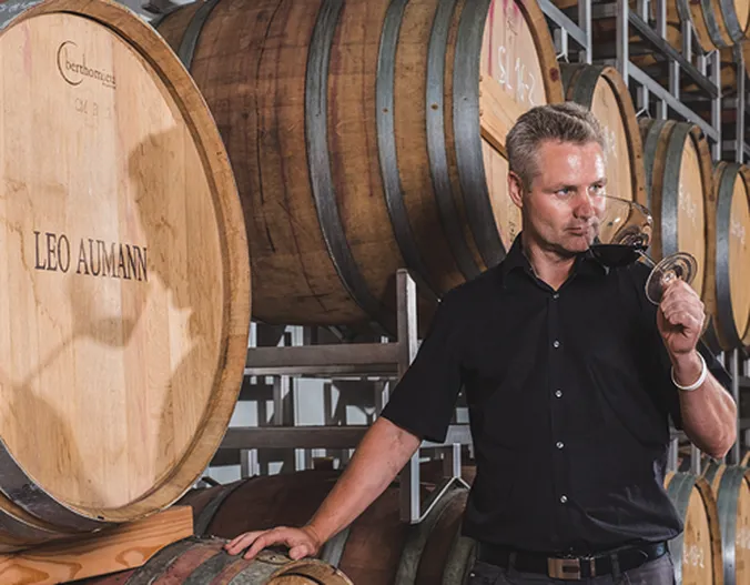 Ein Mann in einem schwarzen Hemd, der aus einem Glas Wein probiert und neben großen Holzfässern in einer Weinkellerei steht.