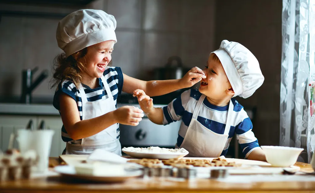 Zwei Kinder mit Kochmützen und Schürzen kochen spielerisch, lachen und veranstalten eine Mehlschlacht in der Küche.