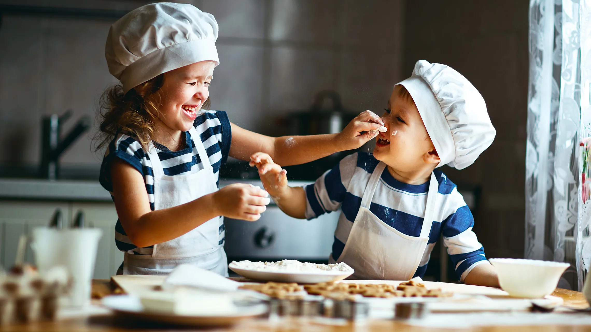 Zwei Kinder mit Kochmützen und Schürzen kochen spielerisch, lachen und veranstalten eine Mehlschlacht in der Küche.