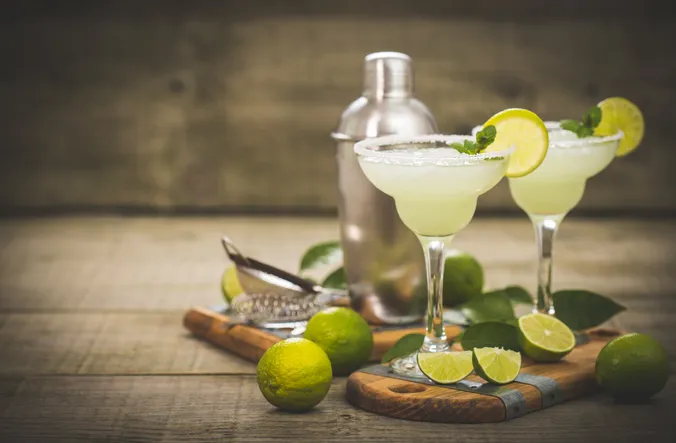 Zwei Margarita-Gläser mit gesalzenem Rand und Limettenscheiben, umgeben von ganzen und in Scheiben geschnittenen Limetten, einem Cocktail-Shaker und einem Sieb auf einer Holzunterlage.