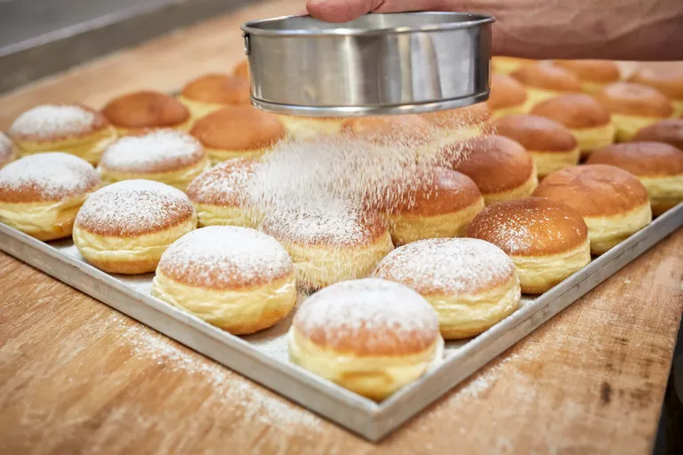 Mehrere frisch gebackene Krapfen werden von einer Person mit Staubzucker angezuckert