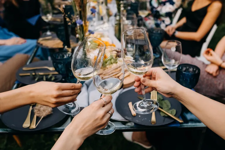 Hände, die bei einer eleganten Veranstaltung im Freien mit Weingläsern anstoßen, im Hintergrund ein gedeckter Tisch mit Geschirr und goldenem Besteck.
