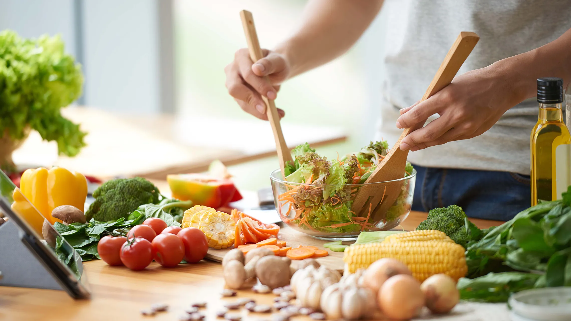 Eine Person, die in einer Schüssel mit Holzbesteck einen frischen Salat anrichtet, umgeben von verschiedenen bunten Gemüsesorten auf einer Küchenarbeitsplatte.