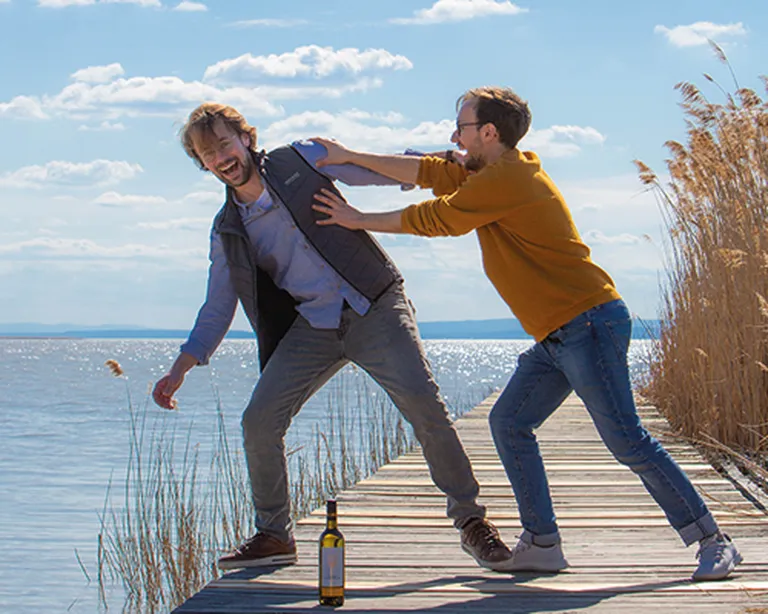 Brüder Nittnaus auf einem Steg am Neusiedlersee mit einer Flasche Wein vor ihnen. Sie lächeln und blödeln herum.