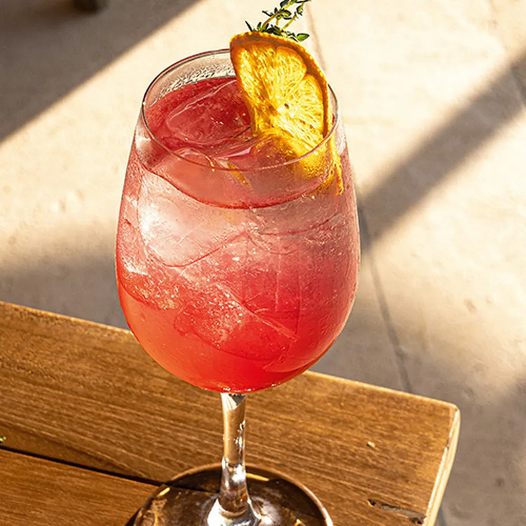 A pink cocktail with ice in a goblet, garnished with an orange slice and a sprig of herbs, on a wooden table in sunlight.