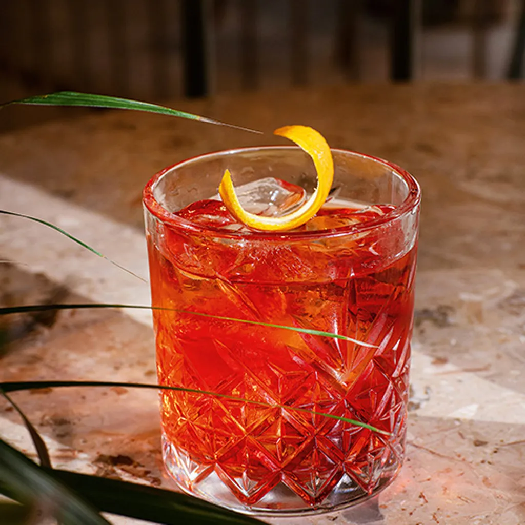 A crystal glass with a vibrant red cocktail, garnished with an orange twist, on a marble table with a plant leaf partially visible.
