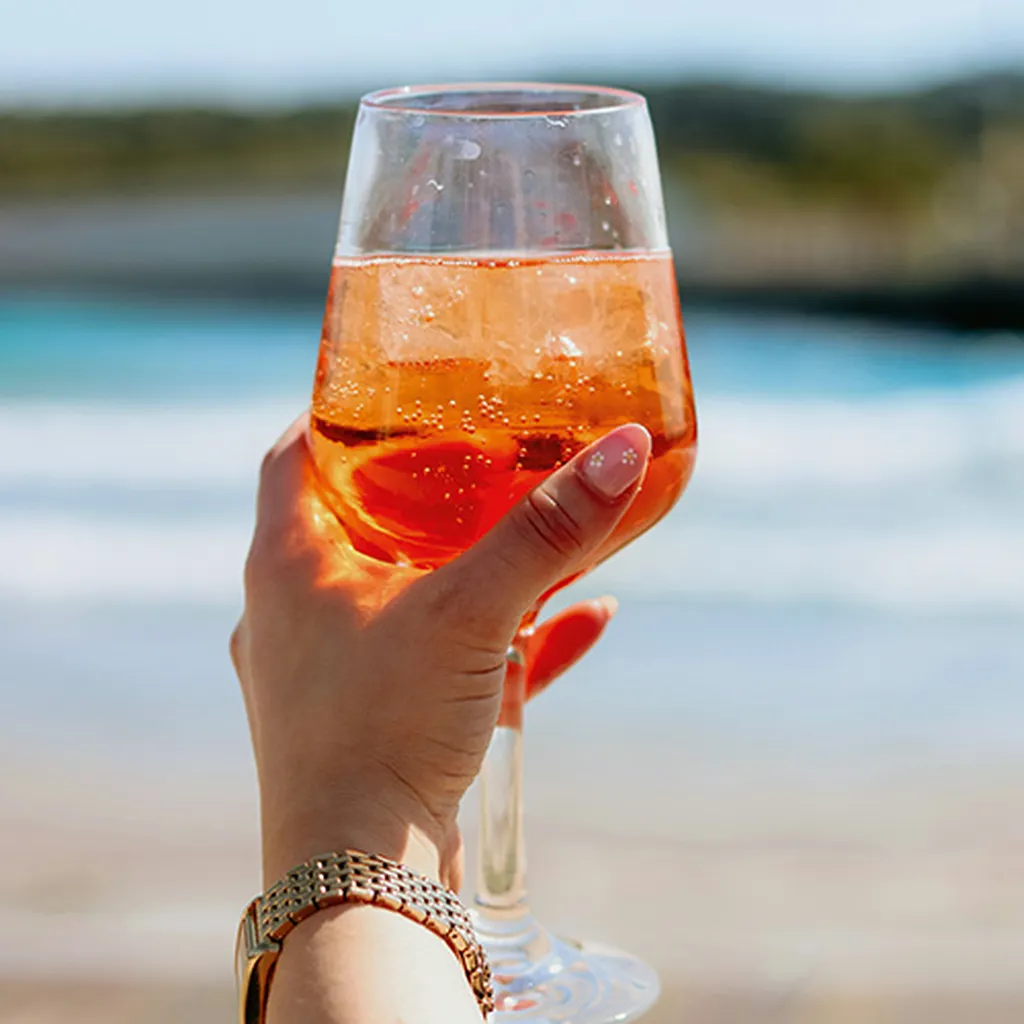 A hand holding a glass of orange cocktail with ice, against a blurred beach background.