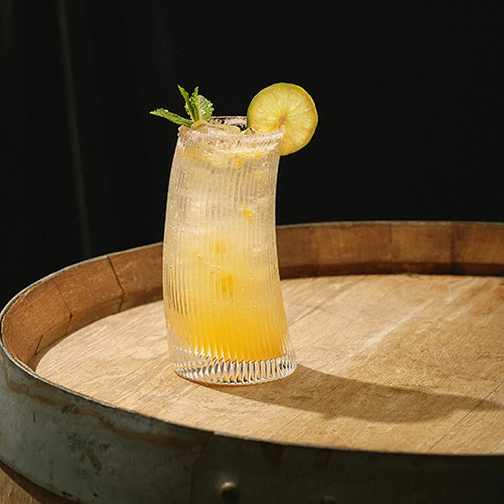 A curved glass with an orange cocktail garnished with a lemon slice and mint sits on a wooden barrel against a dark background.