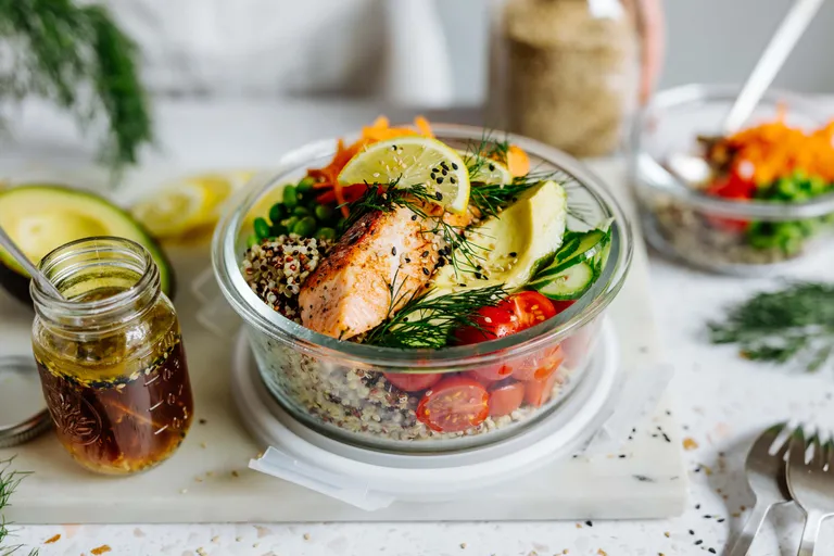 Glasschale mit gegrilltem Lachs, Quinoa, Kirschtomaten, Avocado, Zitronenscheiben und Blattgemüse. Dazu ein Glas Dressing.