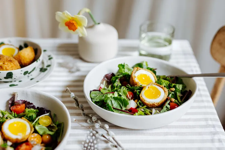 Ein Salat mit halbierten gekochten Eiern auf Blattgemüse, Kirschtomaten und Radieschen in einer weißen Schüssel auf einer gestreiften Tischdecke; daneben steht eine Vase mit einer Blume.