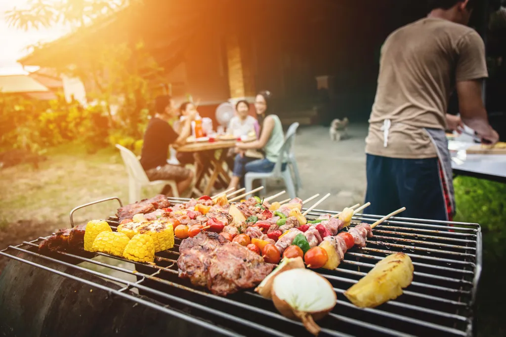 Eine Person, die Spieße, Fleisch, Mais und Zwiebeln auf einem Grill zubereitet, während im Hintergrund Freunde an einem Tisch sitzen.