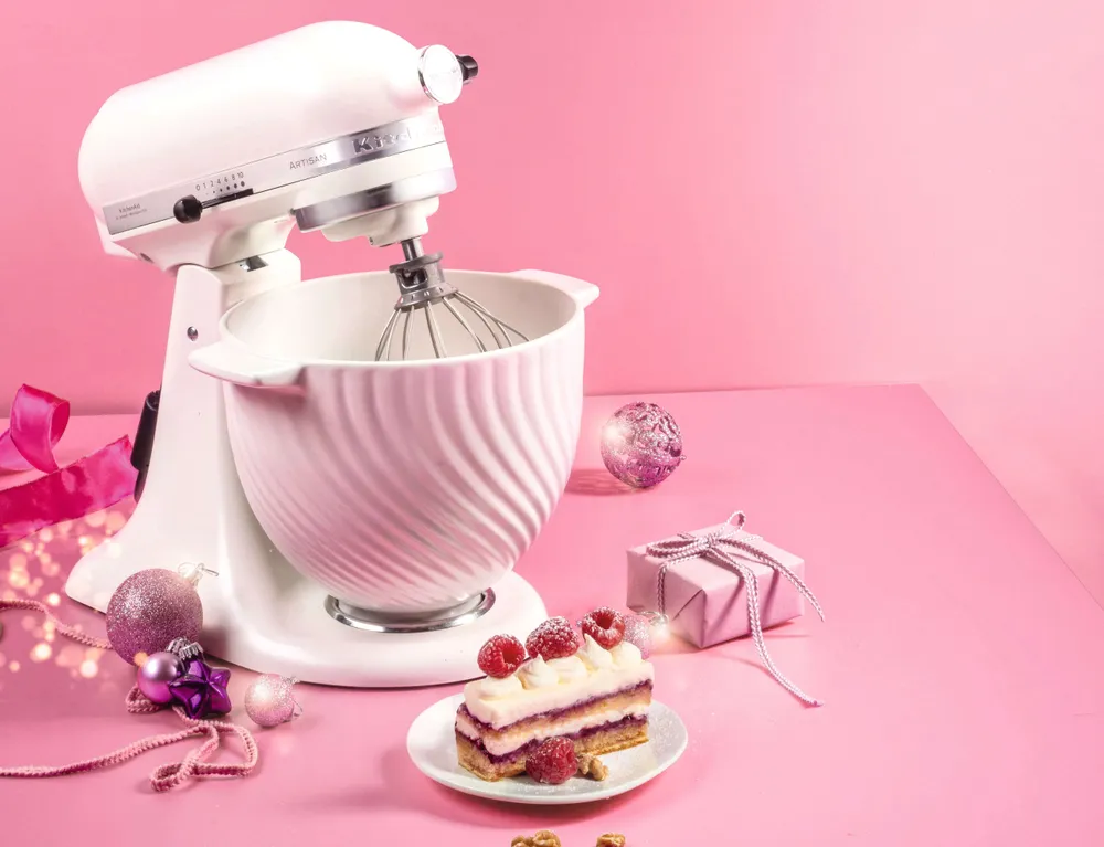 Eine KitchenAid Küchenmaschine auf einem hellrosa Untergrund neben einem StückTorte