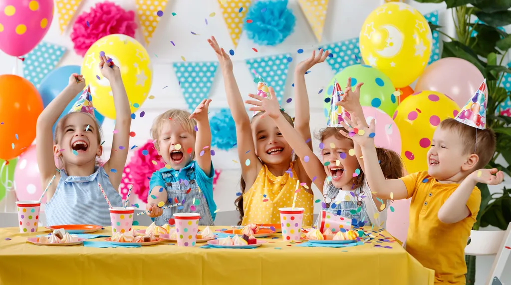 Kinder feiern auf einer bunten Party mit Luftballons, Konfetti und Partyhüten und sitzen an einem Tisch mit Snacks und Getränken.