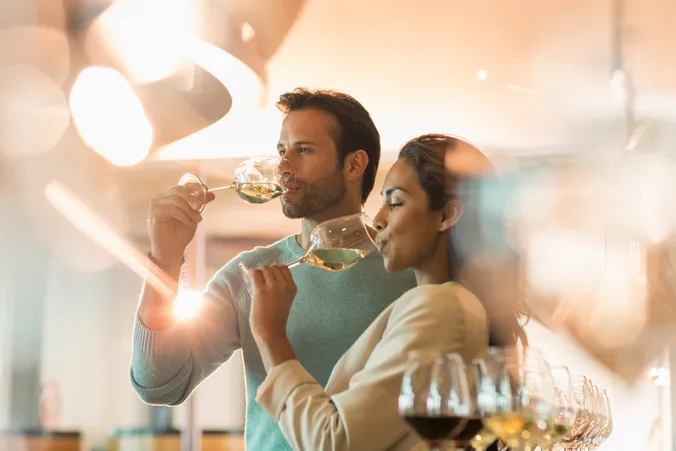 Ein Paar, das in einem sanft beleuchteten Raum Wein verkostet, Gläser mit Weißwein in den Händen hält und von Spiegelungen und unscharfen Elementen im Vordergrund umgeben ist.