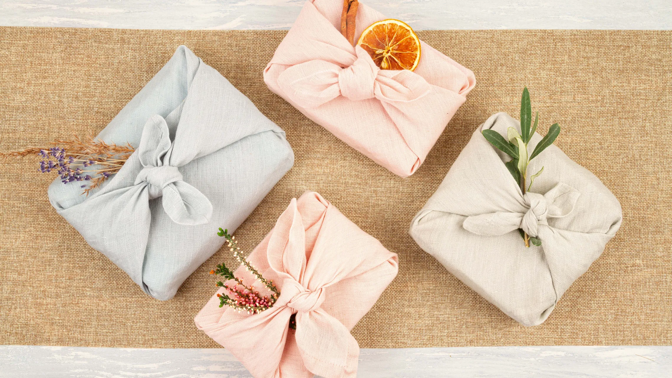 Vier in Stoff verpackte Geschenke, dekoriert mit Naturmaterialien, liegen auf einem Tisch mit Juteläufer