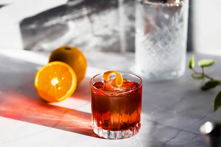 Ein Cocktailglas, gefüllt mit einem rötlichen Getränk und garniert mit einer Orangenschale, daneben eine halbierte Orange und eine ganze Orange auf einer Marmorfläche.