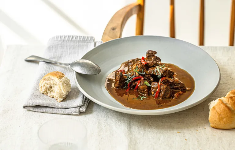 Herzhaftes Rindsgulasch in dunkler Sauce, serviert mit frischen Semmeln auf hellem Tisch.