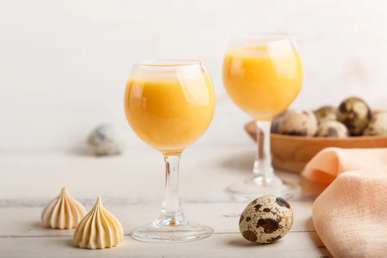 Zwei Gläser Eierlikör mit Wachteleiern, Baisers und einer pastellorangefarbenen Serviette auf einem weißen Tisch.