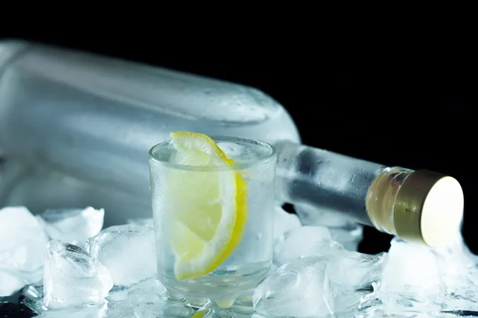 Eine gekühlte Wodkaflasche mit einem vereisten Glas Wodka, garniert mit einer Zitronenscheibe und umgeben von Eiswürfeln.
