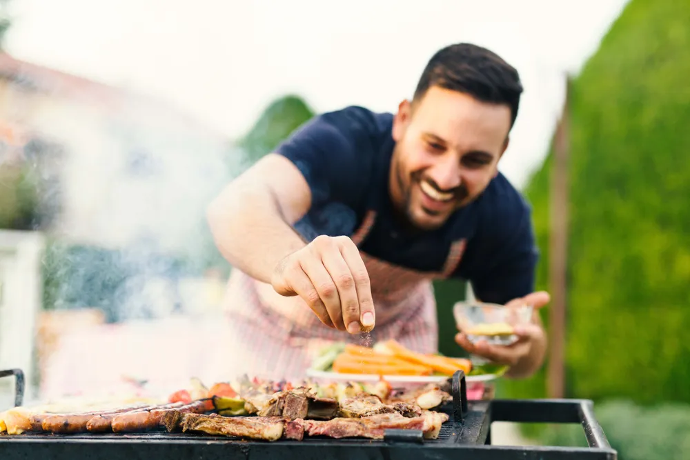 Ein junger Mann steht an einem Grill auf dem sich verschiedene Lebensmittel befinden, die er gerade würzt