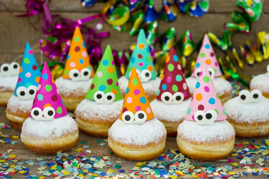 Powdered donuts topped with colorful polka-dot party hats and googly eyes, surrounded by colorful confetti and festive decorations.