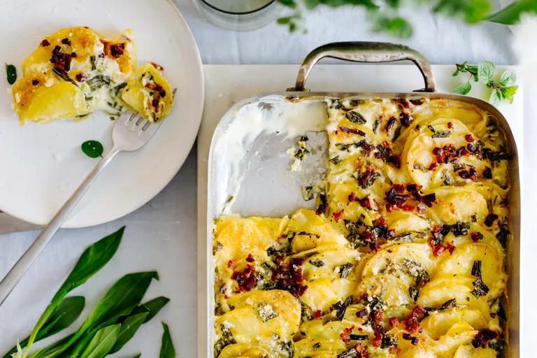 Ofengebackenes Kartoffelgratin mit knusprigem Speck und Kräutern in einer Auflaufform, daneben eine Portion auf einem Teller mit einer Gabel; garniert mit frischem Grünzeug.