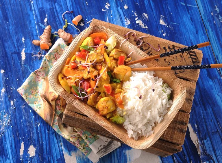 Ein Holztablett mit buntem Hühnercurry und weißem Reis, garniert mit Sprossen, steht auf einem rustikalen blauen Tisch, daneben liegen Essstäbchen.