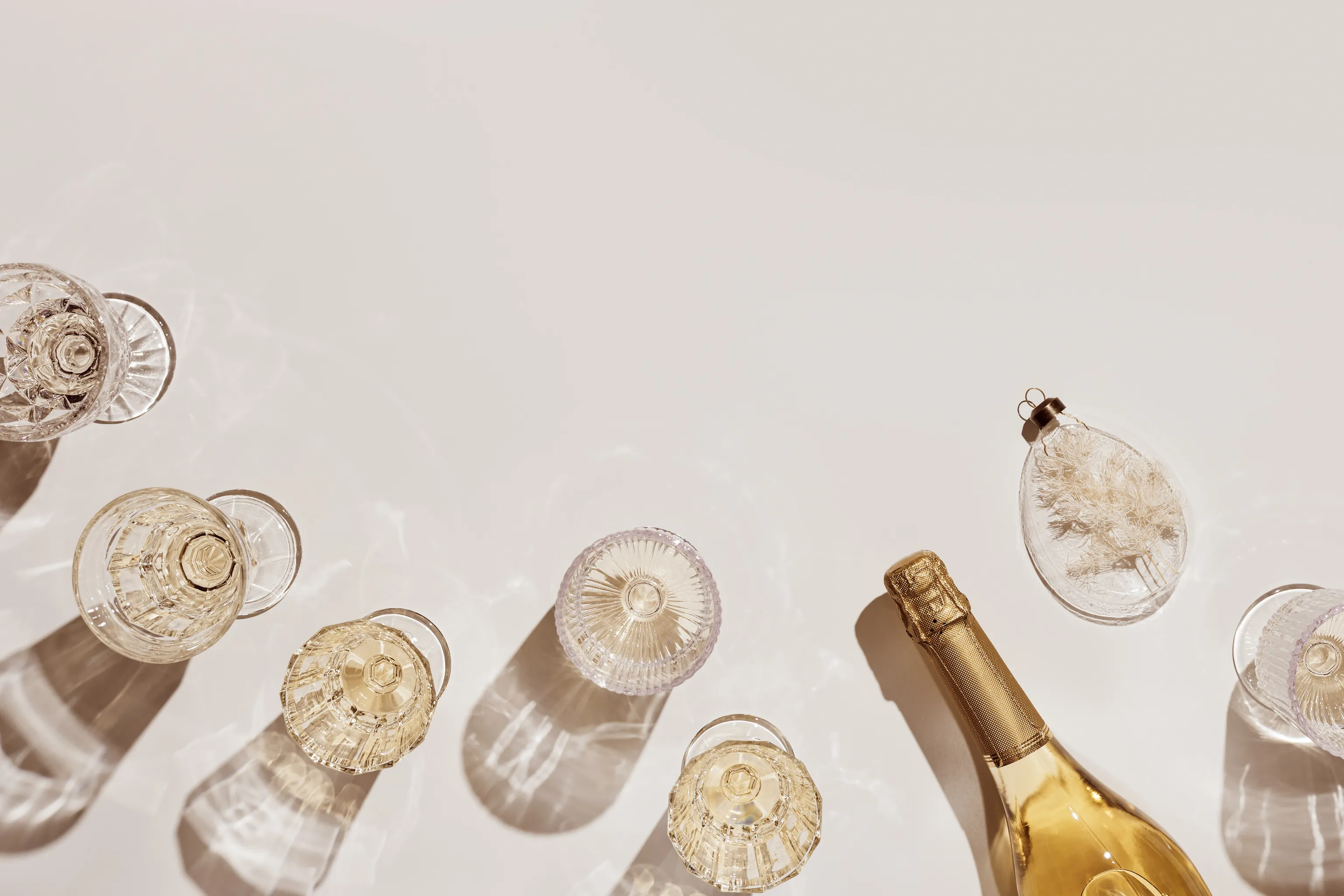 Mehrere leere Vintage-Gläser und eine Flasche Champagner vor einem hellen Hintergrund, die Schatten werfen. Daneben liegt ein zartes dekoratives Ornament.