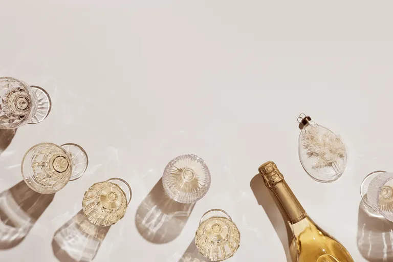 Mehrere leere Vintage-Gläser und eine Flasche Champagner vor einem hellen Hintergrund, die Schatten werfen. Daneben liegt ein zartes dekoratives Ornament.