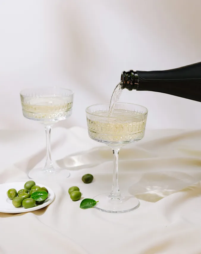 Champagner wird in elegante Kristallgläser eingeschenkt, dazu liegen grüne Oliven und Blätter auf einem kleinen weißen Teller und sind auf einer seidigen Oberfläche verstreut.