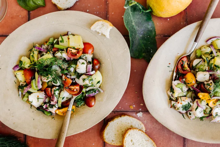 Zwei Schalen mit buntem Meeresfrüchtesalat mit Tomaten, Zwiebeln und Kräutern auf einer Terrakotta-Platte, dazu geschnittenes Brot und eine Zitrone.