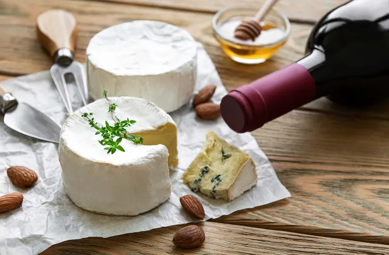 Eine verschlossene Flasche Rotwein liegt neben zwei Weichkäse auf einem hölzernen Tisch